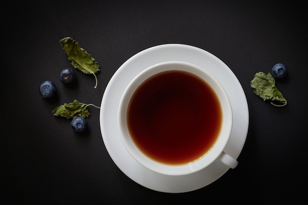 White cup with tea, blueberries and dried mint leaves on a dark