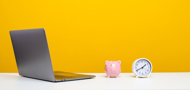 White desk with laptop clock and pink piggy bank yellow background with copy space