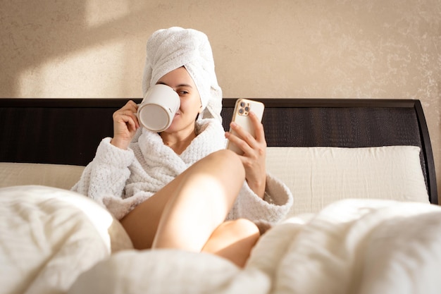 In a white dressing gown with a towel covering her head a woman lies on a bed with a cup and a phone in her hand