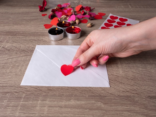 White envelope next to candles with red rose petals on the table and a woman's hand holding a heart