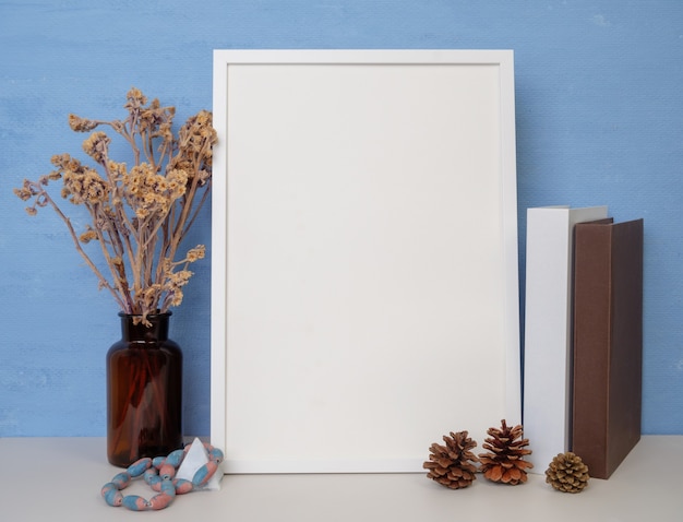 White frame mockup for design or text, books,dried flower in glass jar,pine cones and antique stone sitting on a wooden table blue wall background