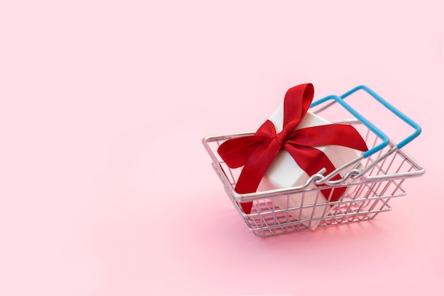 White gift box with red bow in supermarket basket on pink background