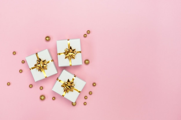 White gift boxes with golden bows and golden decorations on pastel pink background. Minimal styled holiday card. Flat lay, top view, copy space.