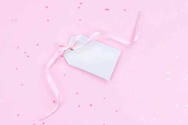 White gift tag with pink ribbon on pink background with heart shape confetti