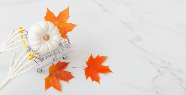 White halloween pumpkin in little grocery trolley with skeleton arms and maple leaves on marble back