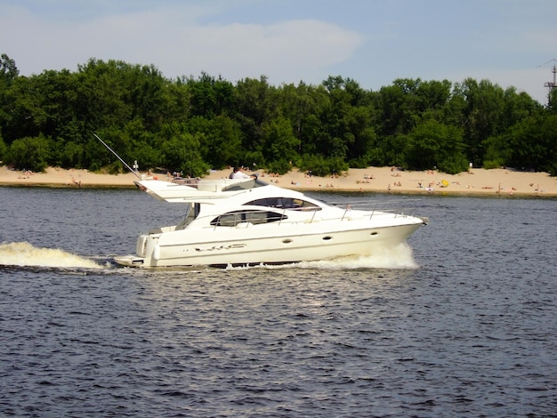 White high-speed yacht sails on the background of the beach with sunbathing people and trees