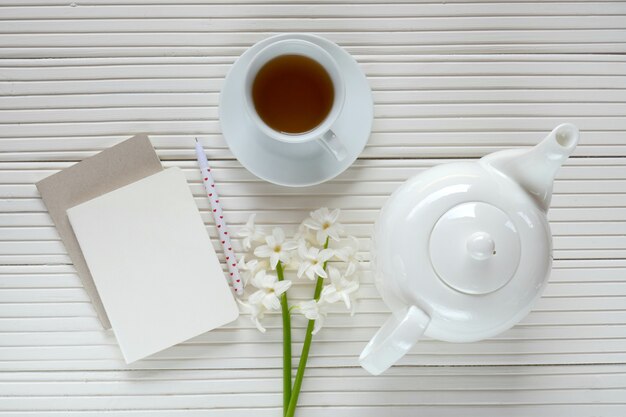 Photo white hyacinth flower, cup of tea, white teapot and blank notebook on a white wooden board background