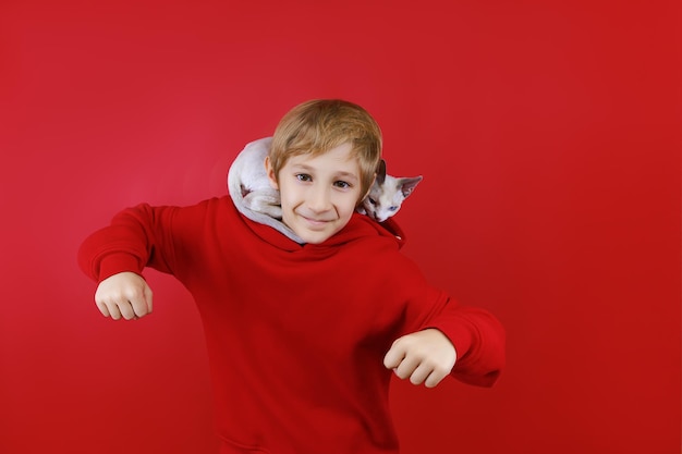 A white kitten climbed into the hood of a boy who stands on a red background