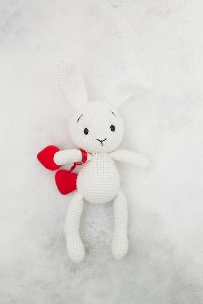 A white knitted bunny holds red hearts on a gray background