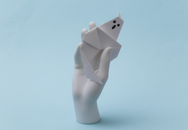 White mannequin hand holding paper origami ghost on blue background. Halloween concept