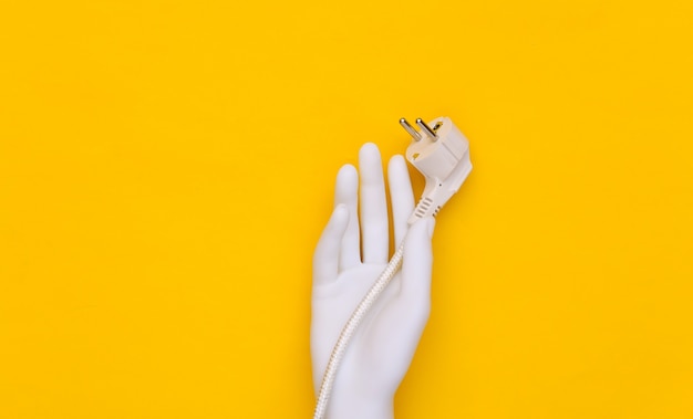 White mannequin hand holds electric plug with cord on yellow background.