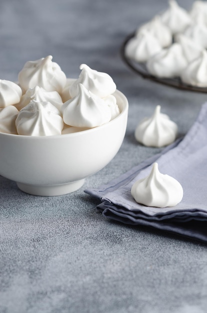 White meringue cookies in a white bowl with a light blue napkin, on grey background.