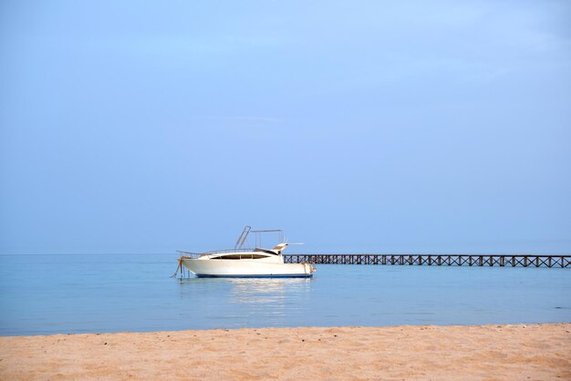 White motor boat floats in ocean water at long pier bridge under bright blue sky view from sandy beach Summer traveling concept