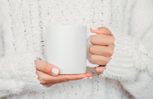 White mug mockup. Girl is holding white 11 oz ceramic cup in hands, wears cozy knitted sweater. Front view, copy space.