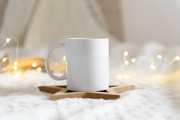 White mug mockup with blank copy space. Coffee cup on a beige knited background with opened book and christmas garland lights, cozy winter day at hugge home