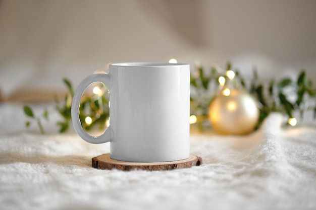 White mug mockup with blank copy space. Coffee cup on a beige knited background with opened book and christmas garland lights, cozy winter day at hugge home