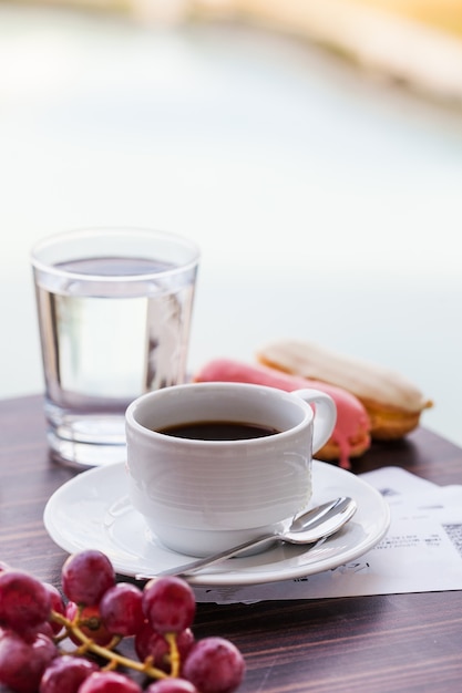 White mug with black coffee with eclairs and a glass of drinking water