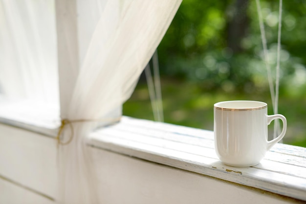 White mug with tea or coffee on a white wooden table on summer terrace in green garden background