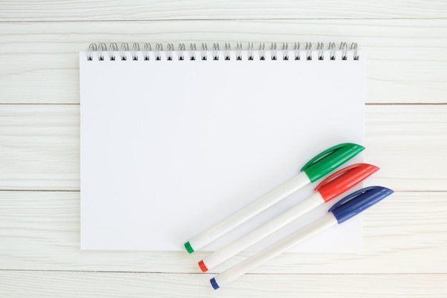 White notebook on metal spiral and three green red and blue ballpoint pens on wooden table