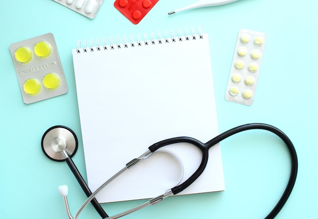 White notepad that lies next to the stethoscope and pills on a blue background. Medical concept