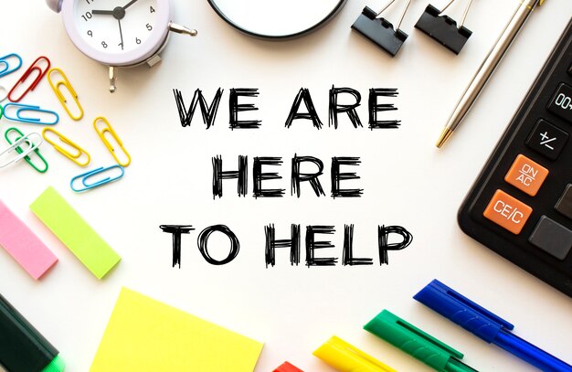 White office desk table with calculator, magnifying glass, colored pens and other stationery. Text on the WE ARE HERE TO HELP. View from above. Business concept.