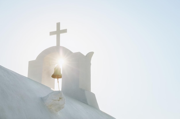 White Orthodox Church belfry with cross and bell in sunshine on sky background Santorini island Greece