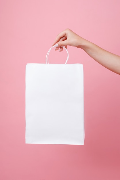 White paper bag under the logo in the hands of the girl on a pink space. Shopping mock up holding