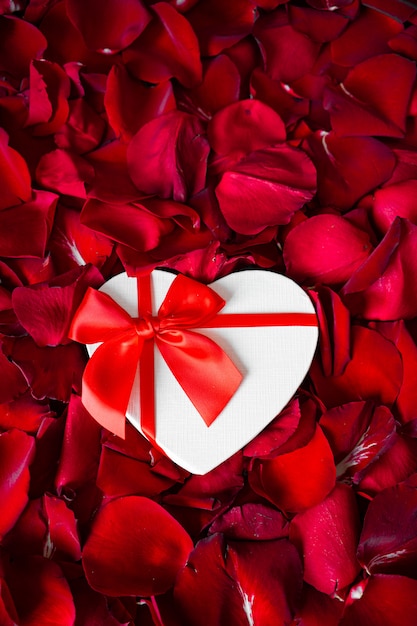 White paper heart with red bow on background of red rose petals