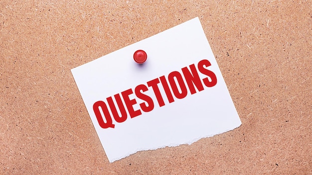 White paper with the text QUESTIONS is attached to the wooden background with a red button