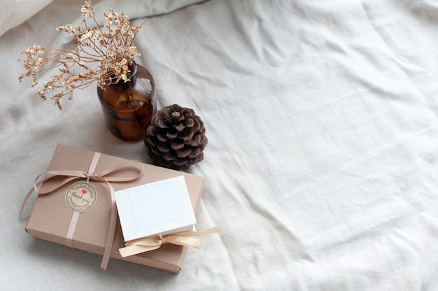 A white picture frame placed in a brown gift box surrounded by a box of small diamond rings tied with cream-colored pine cones dried flowers in a brown glass bottle.