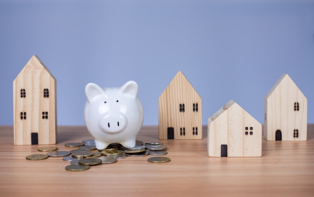 A white piggy bank is placed on a pile of coins. And beside it is a wooden model house on a white background. which are all on a wooden table