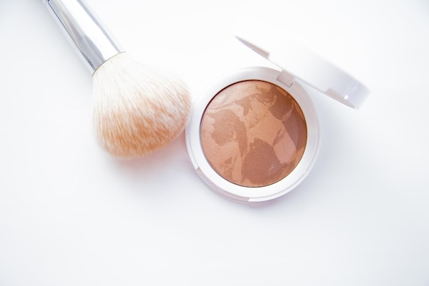 A white pillbox with powder and bronzer lies on a white background next to it lies a brush for blush