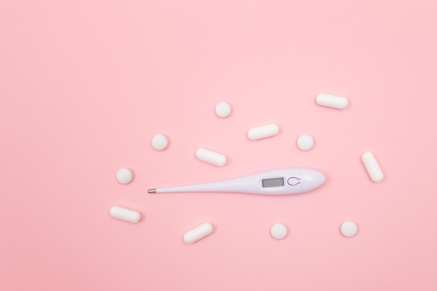 White pills or tablets with electronic thermometer on pink background