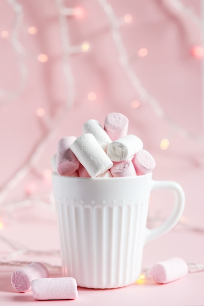 White and pink marshmallows in a white cup on a pink background. Glowing lights in the background