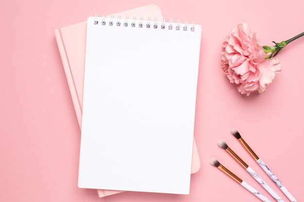 White and pink notebook with carnation flower and make up brushes on a pink background