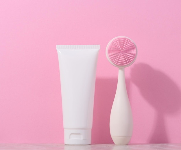 White plastic tube and a massage brush for facial cleansing on a pink background items for cosmetic procedures