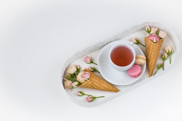 in a white plate a cup of tea, macarons in waffle cones and pink roses