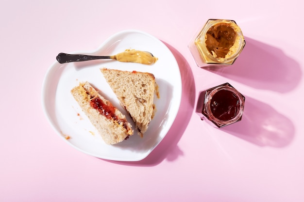 White plate in shape of heart with sandwiches with peanut butter and strawberry jelly on pink background. Top view