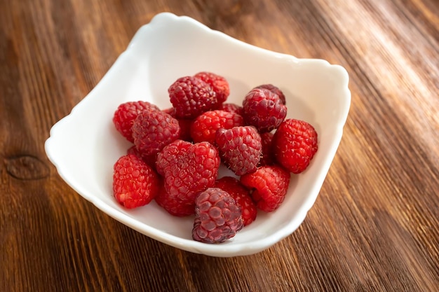 In a white plate which stands on a wooden table juicy fresh raspberries on which the rays of the sun fall