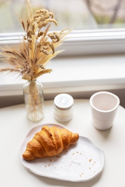 A white plate on which there is a croissant and a glass of clay with an inscription handmade are near the window on the table