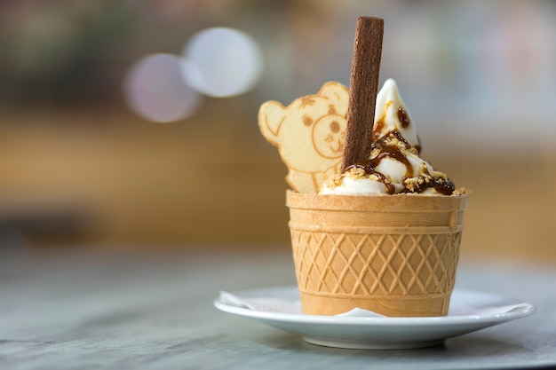 White plate with ice cream dessert in wafer cup with chocolate cookies and creative decoration topping on blurred colorful interior.