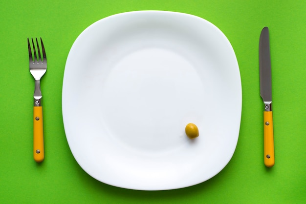 White plate with one olive and cutlery on green tablecloth