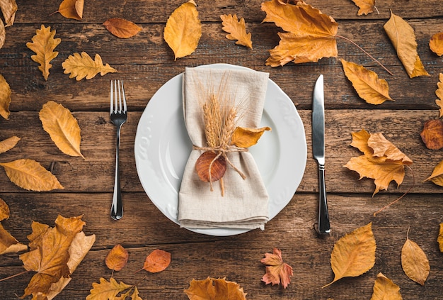 A white plate with a serving napkin and cutlery on a wooden background with autumn leaves