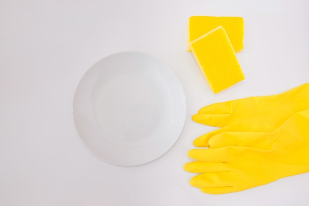 White plate with yellow gloves and yellow washcloths on a white background. Copy space. View from above.