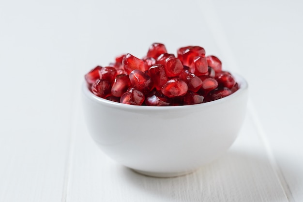 White porcelain Cup with ripe pomegranate seeds on a white wooden table. Vegetarian food.