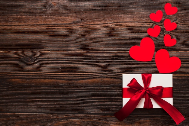 White present with a red ribbon and paper hearts isolated on a dark wooden background. Top side view of a celebrating warm flatlay. Valentines day and Christmas concept. Copyspace.