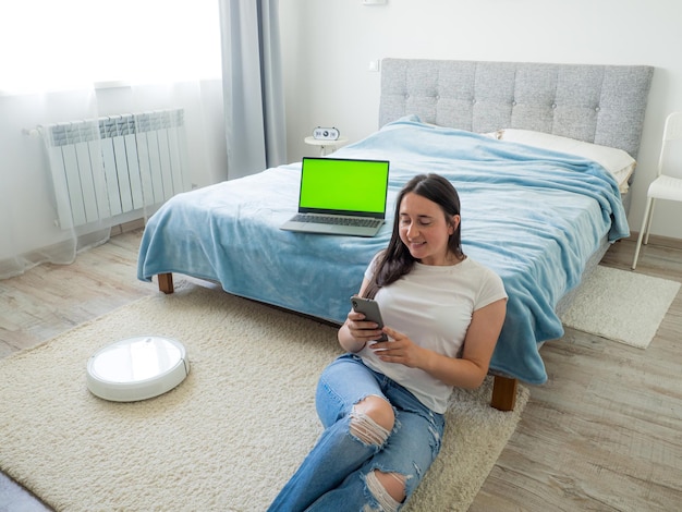 White robotic vacuum cleaner cleaning the floor while woman using smartphone