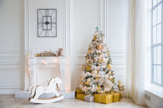 White room interior with New Year tree decorated, present boxes and artificial fireplace