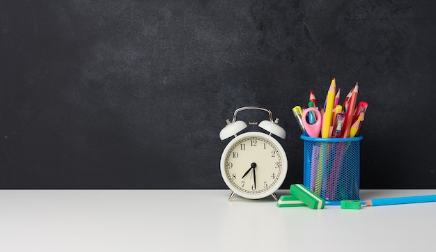 White round alarm clock and a metal glass with pens, pencils and felt-tip pens on the background of an empty black chalk board, copy space