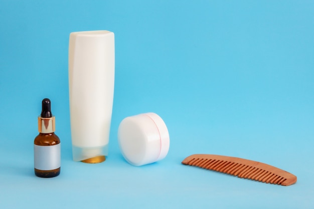 White round cream jar, pipette jar, wooden comb, large white bottle are on a blue sky background, front view